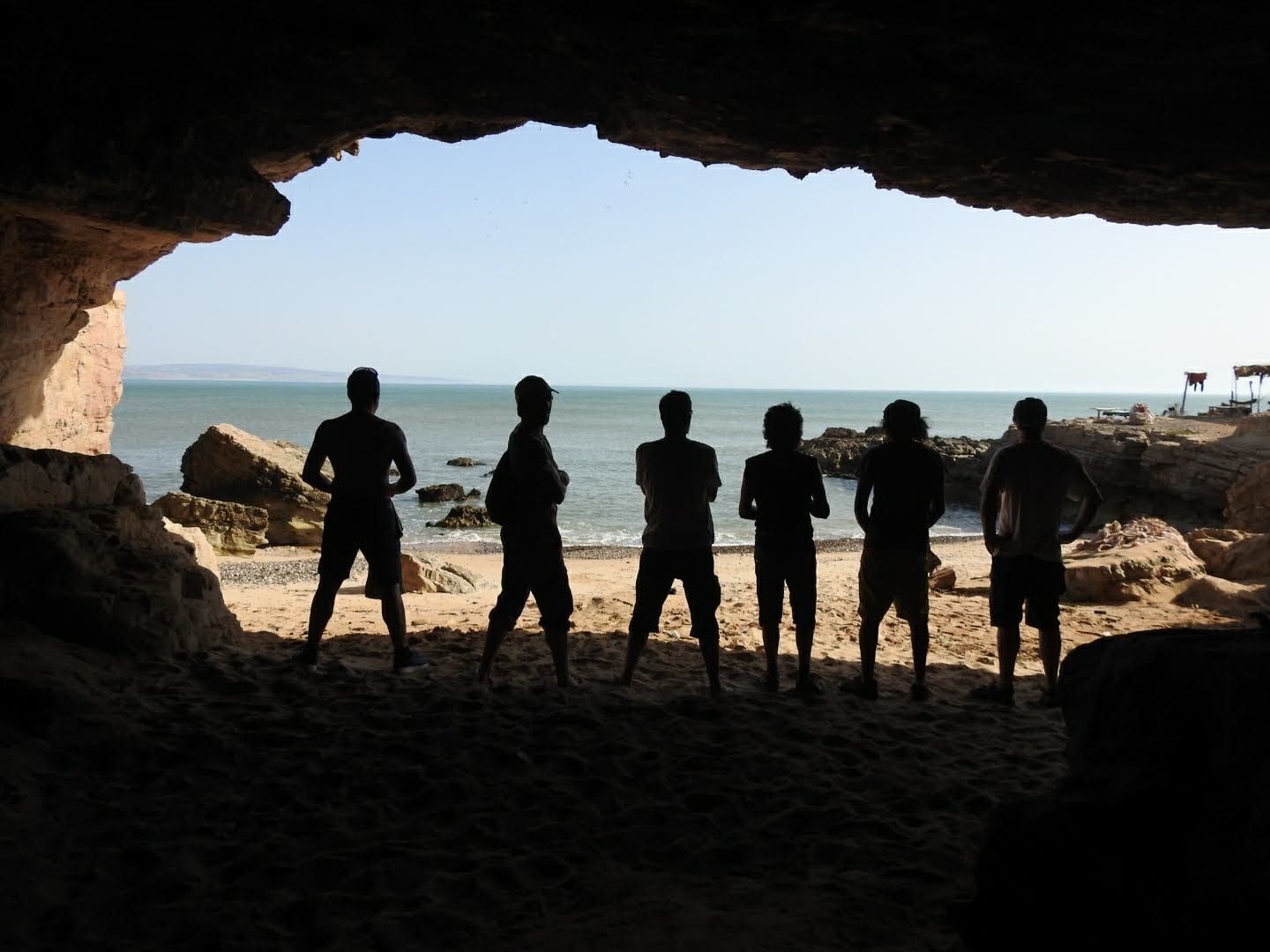 Six silhouettes se découpent dans l’ouverture d’une grotte marine, face à l’océan et à une plage de galets