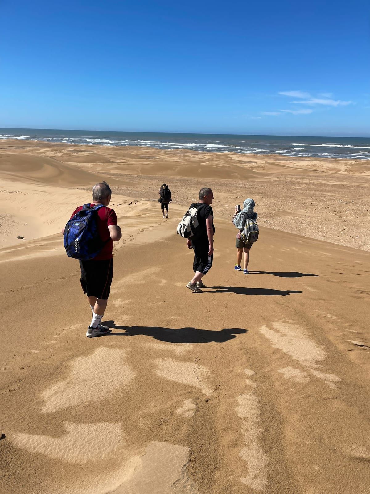 Quatre randonneurs sac au dos progressent sur la crête d’une dune, vus de dos