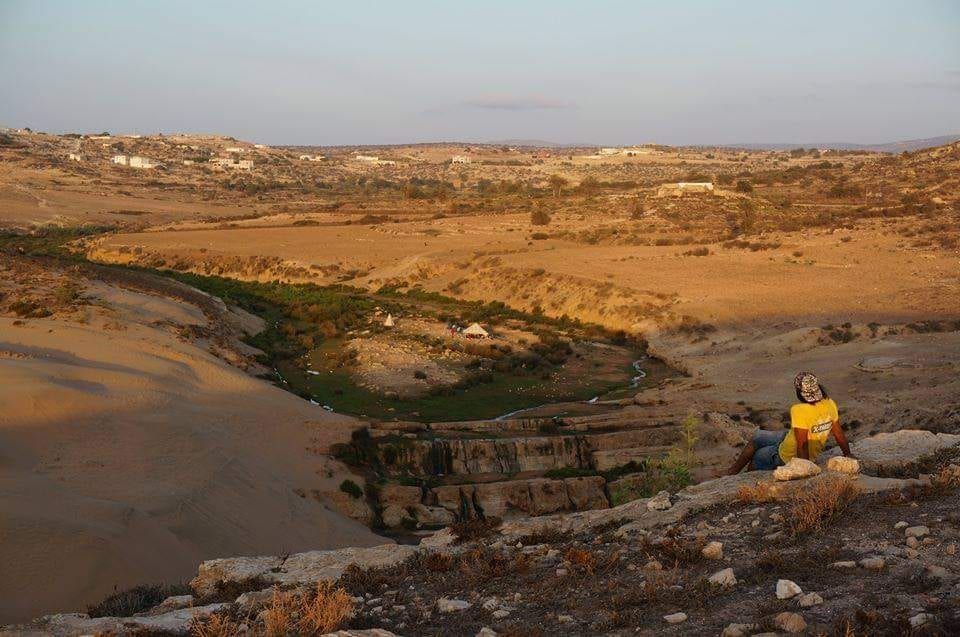 Au couchant, une personne assise sur un promontoire contemple une vallée verte creusée par l’oued, une tente de bivouac au loin