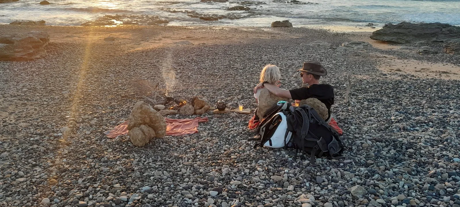 Deux marcheurs assis près d’un feu, sur la plage de galets, au soleil couchant