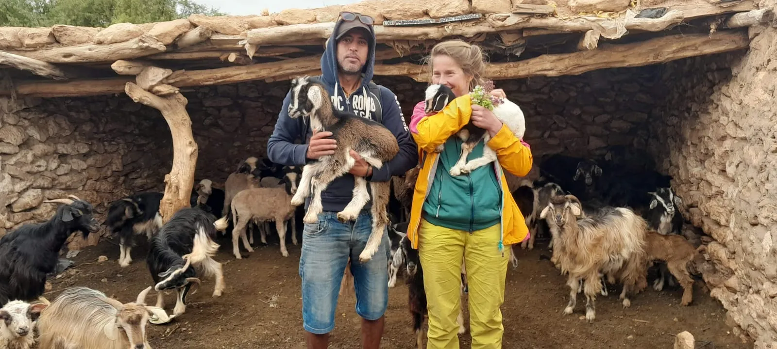Deux visiteurs tiennent chacun un chevreau sous le toit de branches de la bergerie