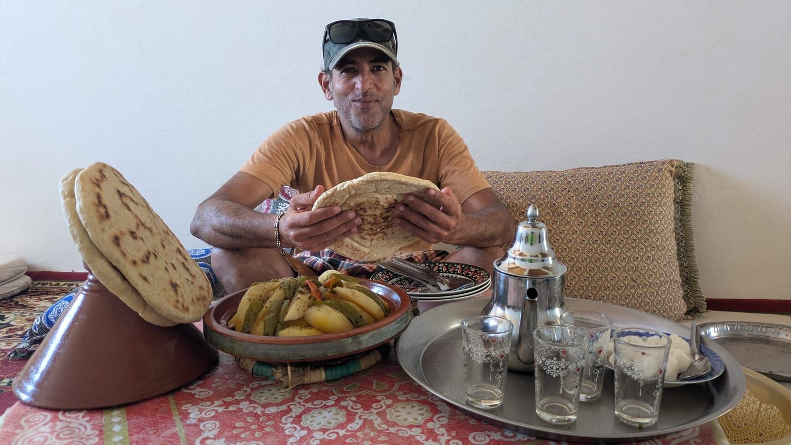Kabir assis devant un tajine, tenant un pain rond ouvert à deux mains, théière et verres posés sur le plateau