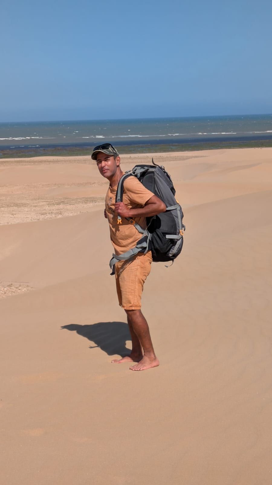 Kabir marche sur une longue plage de sable, sac à dos chargé, l’océan derrière lui