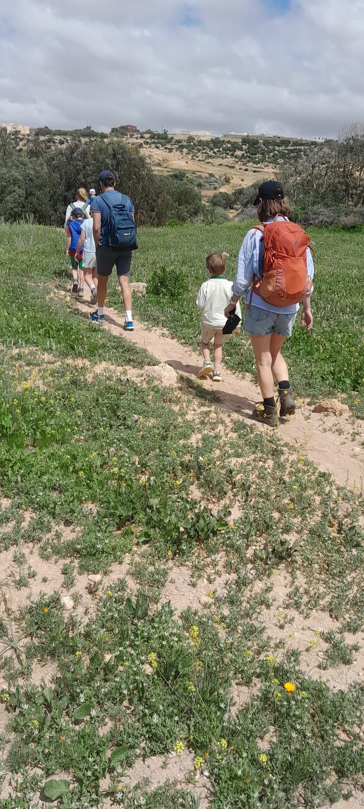 Une famille marche en file sur un sentier de terre, vue de dos, entre les herbes hautes